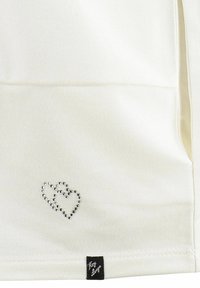 Top in tessuto bianco con un motivo a cuore tempestato di strass argentati e un piccolo etichetta nera sull'orlo. Texture setosa, design semplice.