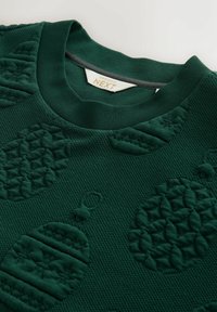 Pull en tricot vert foncé avec des motifs de décorations de Noël en relief et une étiquette "Christmas at Next" à l'intérieur du col.