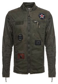 Olivgrüne Zip-Jacke mit Aufnähern: Stern, "BE EDGY", "BORN READY", EU-Flagge und rotes Design mit einer schwarzen Hand.