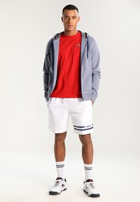 Mann med en grå glidelåshette over en rød t-skjorte, hvite shorts med blå striper, og hvite sneakers med røde detaljer.