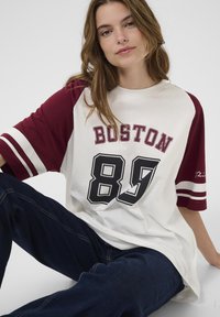 Bílý tričko s krátkým rukávem a maroon raglánovými rukávy, s nápisem "BOSTON" a "89" v černé barvě. Měkká bavlněná směs s vodorovnými bílými pruhy.