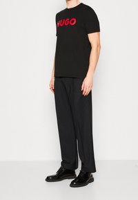 Schwarzes T-Shirt mit kurzen Ärmeln und rotem "HUGO"-Logo, getragen mit lockeren schwarzen Hosen und glänzenden schwarzen Schnürschuhen. Einfaches, modernes Design.