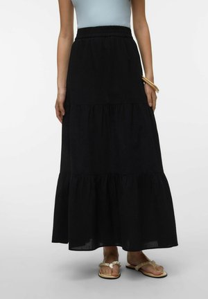 Femme portant une longue jupe noire à volants, un top sans manches bleu clair, des sandales beiges et des bracelets en bois au poignet.