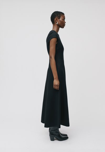 Robe noire longueur genoux à manches courtes, en tissu lisse. Assortie avec des bottes chelsea noires à motifs et des boucles d'oreilles argentées simples.