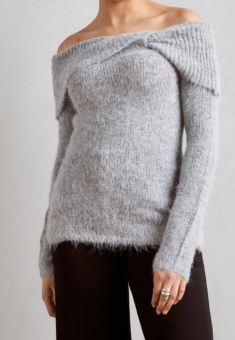 Femme portant un pull en maille duveteuse gris clair, épaules dénudées, et un pantalon foncé, montrant de longues manches et un détail de col double.