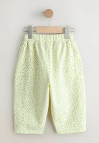 Ljusgula byxor med ett diskret grönt prickmönster, mjukt tyg, elastisk midjeband och kort längd. Visas på en klämma.
