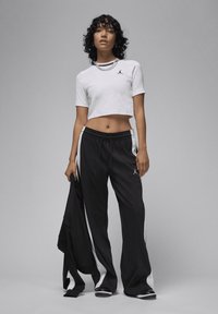 White cropped t-shirt with a black logo, paired with black track pants featuring white side stripes and a drawstring waist. User holding a jacket.