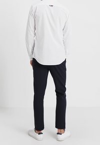 Camisa blanca de manga larga con dobladillo curvado, combinada con pantalones oscuros y zapatillas blancas, vista desde atrás. Diseño minimalista.