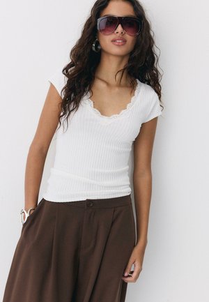 Frau mit großen dunklen Sonnenbrillen, silbernen Creolen, weißem, geripptem Kurzarmshirt mit Spitzenbesatz und hoch taillierter brauner Hose.