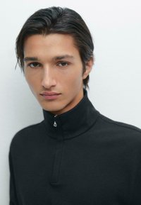 Zwarte hoge hals pullover met een halve ritssluiting, gemaakt van zacht materiaal. Het heeft een gladde textuur en een minimalistisch ontwerp zonder zichtbare patronen of accenten.