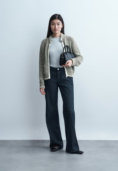 Νεαρή γυναίκα με μακριά σκουρόχρωμα μαλλιά, φορούσα γκρι πουλόβερ, υφασμάτινο μπουφάν με φερμουάρ, φαρδιά σκούρα τζιν παντελόνια, μαύρα παπούτσια, κρατώντας μια μικρή μαύρη τσάντα χειρός.