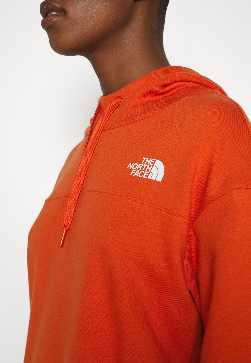 Gros plan sur une personne portant un sweat à capuche orange vif de The North Face avec des cordons de serrage sur la capuche, sur un fond blanc uni.