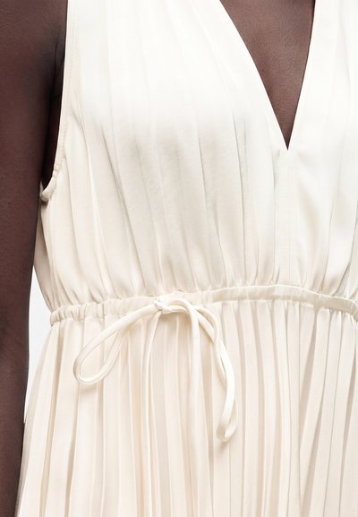 Gros plan d’une personne à la peau foncée portant une robe blanche sans manches, plissée, avec un décolleté en V profond et une ceinture nouée à la taille.