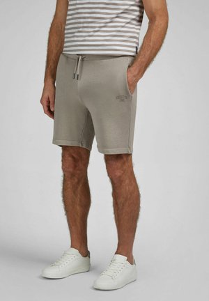 Man draagt beige short met trekkoord en logo "HECHTER 1962", gestreept shirt en witte sneakers, staand tegen een effen achtergrond.