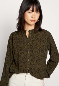 Camisa verde com padrão de manchas pretas, frente com botões, gola redonda e mangas longas, combinada com calças de cor clara.