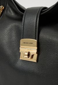 Sac à main en cuir noir avec une texture lisse, des finitions dorées et un fermoir carré avec un logo imprimé. Double anse pour un transport facile.