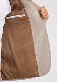 Beiger Blazer mit einem strukturierten Außenstoff und glattem braunem Futter. Verfügt über eine Tasche mit weißem Besatz innen. Eine Hand hält die Jacke offen.
