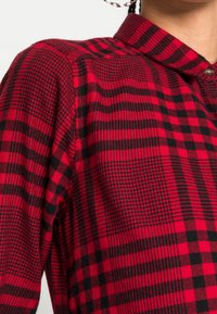 Red and black checkered shirt with a collar, featuring a mix of plaid and houndstooth patterns in a textured fabric. Button detail visible.