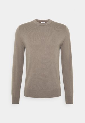 Henkilö, jolla on musta takki, mustat housut, mustat kengät ja beige toppi, pitelee mustaa taiteltua laukkua valkoisen taustan edessä.
