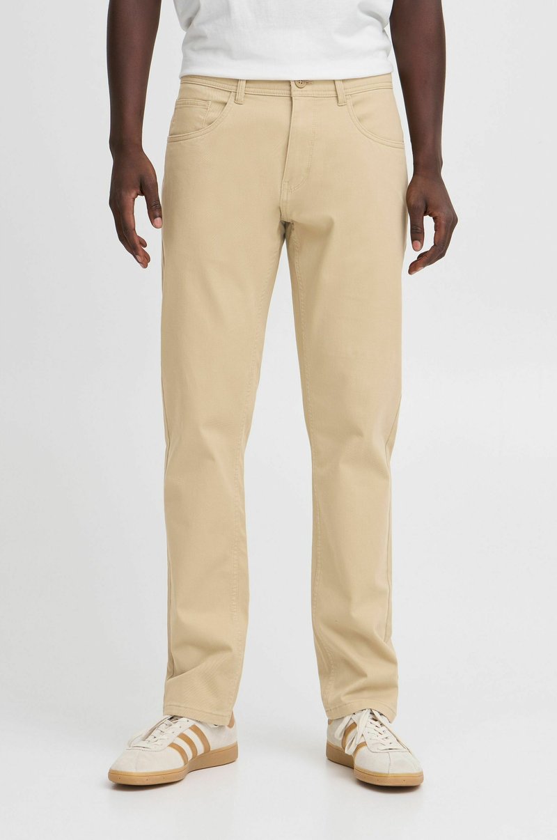 Pantalons en mélange de coton beige avec une coupe droite, design à cinq poches et détails de couture visibles, associés à des baskets blanches.