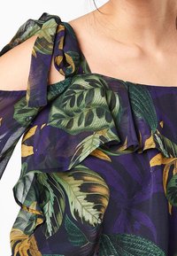 Tessuto a stampa floreale con grandi foglie verdi e gialle su uno sfondo viola scuro. Collo arricciato con un nodo regolabile su una spalla.