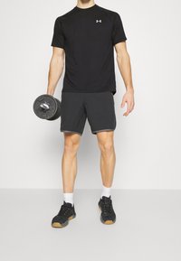 Svart idrottströja, grå shorts med nederkantaccent, håller i en svart hantel, bär svarta sneakers och vita strumpor. Neutral bakgrund.