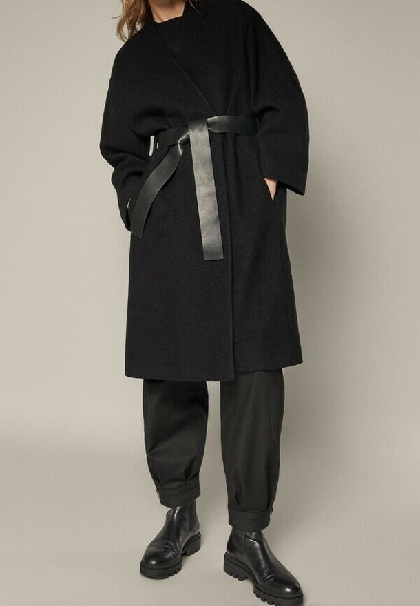 Sort uldblanding frakke med bælte, brede ærmer, sidelommer, parret med sorte tapered bukser og ankelstøvler, sat mod en neutral baggrund.