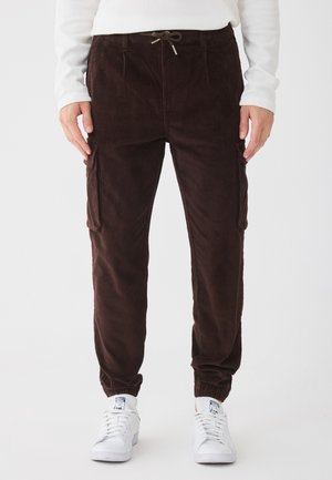 Brune kordfløyels joggebukser med side cargo lommer, elastiske mansjetter og snøring i livet, kombinert med hvite joggesko.