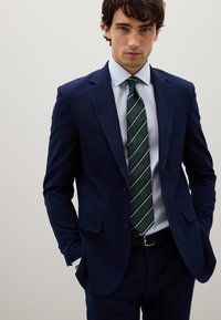 Abito blu navy in tessuto texturizzato, abbinato a una camicia azzurra e una cravatta a righe verdi. Design aderente con tasche frontali e cintura.
