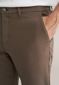 Pantaloni maro dintr-un material neted, cu un buzunar lateral, închidere cu buton și un design slim. Fără modele sau detalii hardware vizibile.