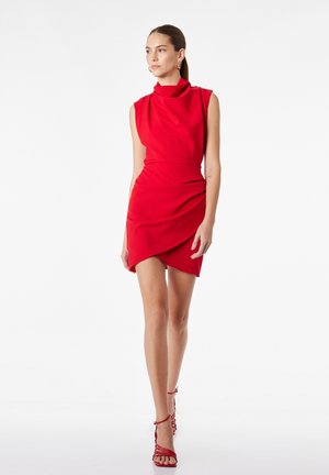 Mujer de pie con un vestido mini rojo sin mangas con un cuello alto drapeado y sandalias de tacón rojas con tiras.