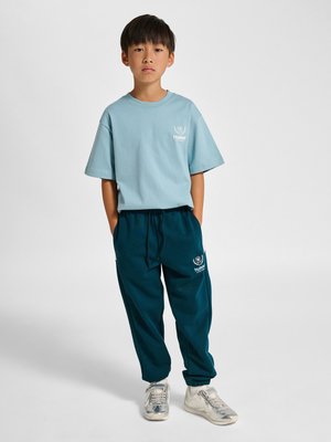 Junge trägt hellblaues T-Shirt und dunkelblaue Jogginghose mit weißen Logos, steht mit Händen in den Taschen vor einfarbigem Hintergrund.