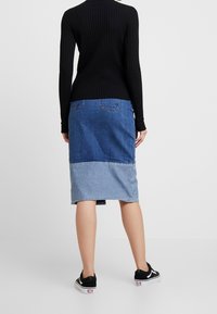 Femme debout, portant un pull côtelé noir, une jupe midi en denim bicolore bleue, et des baskets basses noires avec des semelles blanches.