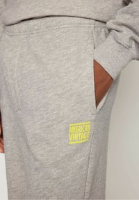 Sive trenirke iz mehkega materiala, z stranskim žepom in svetlo rumenim logotipom "AMERICAN VINTAGE" na sprednji strani.