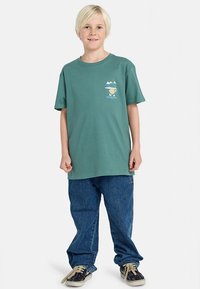 Grøn bomuld T-shirt med korte ærmer, med et grafisk tryk på venstre bryst. Bæres med løstsiddende blå cowboybukser og sneakers.