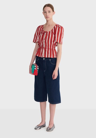 Jeune femme portant un chemisier rayé rouge et blanc, un jean large cropped foncé, des ballerines argentées, tenant une petite pochette colorée.