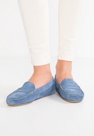 Pieds portant des mocassins en suède bleu clair assortis à un pantalon blanc ajusté sur fond blanc.