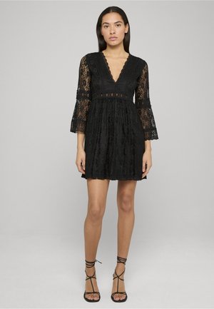 Sort blondekjole med v-udskæring, klokkeærmer og lagdelt skørt. Tekstureret stof med blomstermønster og indsnævret talje. Sorte sandaler med remme.