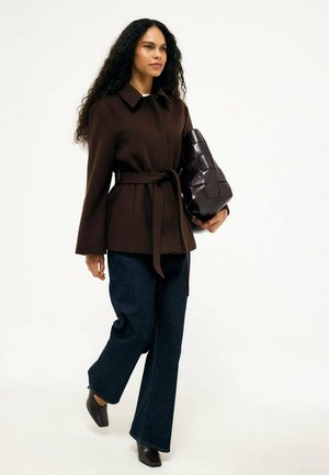 Mujer con cabello largo y rizado caminando, vistiendo un abrigo marrón con cinturón, vaqueros azul oscuro, zapatos negros y llevando un gran bolso negro tejido.