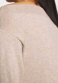 Maglione beige con superficie texturizzata e scollatura ampia, caratterizzato da dettagli a coste lungo le spalle e i polsini.