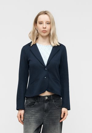 Blondína s rovnými vlasmi, ktorá má strieborné náušnice, tmavomodrý blejzer cez biele tričko s vzorom a vyblednuté čierne rifle na jednoduchom pozadí.