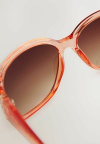 Lunettes de soleil surdimensionnées avec des verres dégradés marron et une monture en acétate translucide corail-orangé. Présente des bords arrondis et des détails matériels subtils.