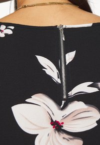 Tissu noir avec un grand motif floral ; présente une fermeture éclair argentée au dos. Texture lisse avec des couleurs contrastées, y compris le blanc et le rouge.