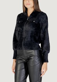 Veste zippée en fausse fourrure noire avec un col, deux poches poitrine et des bords côtelés, associée à un pantalon en cuir noir.