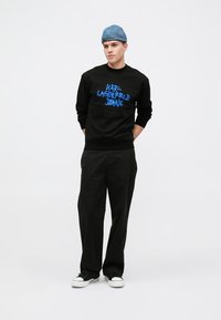 Svart hettegenser med blå grafisk tekst "KARL LAGERFELD JEANS," kombinert med svarte bukser og hvite joggesko. Tilbehør inkluderer en blå caps.