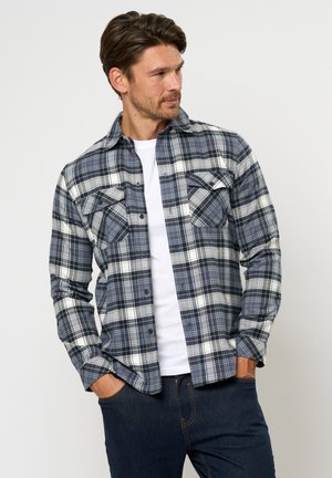 Man som bär en blå och vit rutig skjorta över en vit T-shirt, med händerna i fickorna på mörka jeans, tittar åt sidan.