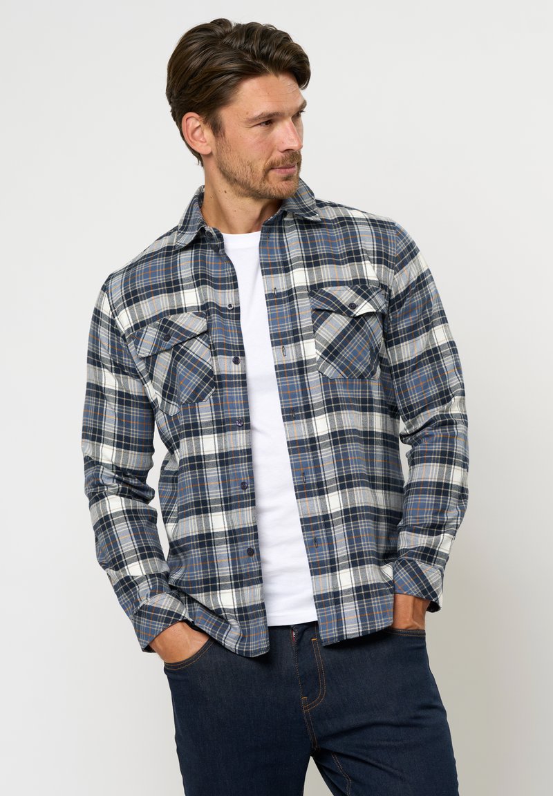 Mees, kes kannab sinise-valge ruudulise trukiga särgi ja valget T-särki, käed tumedates teksataskutes, vaatab küljele.