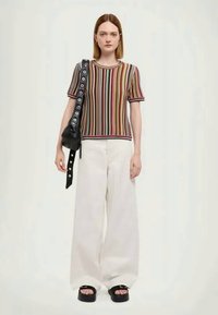 Kurzärmliges Oberteil mit bunten Streifen, kombiniert mit weißen weiten Hosen. Das Model hält eine schwarze Tasche mit runden Beschlägen. Schwarze Plateausandalen.