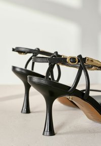 Mustat korot sandaalit kiiltävällä pinnalla, ohuilla remmeillä, kultaisilla yksityiskohdilla ja hoikalla stiletto-korkolla. Sujuva pinta, minimalistinen muotoilu.