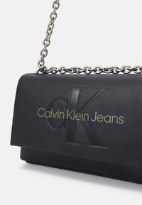 Bolso de hombro de cuero sintético negro con una correa de cadena plateada, que presenta un logotipo 'CK' en relieve y texto verde que dice "Calvin Klein Jeans."